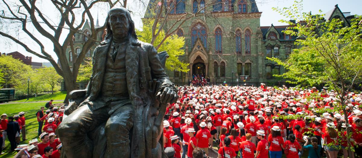 A view of the Benjanmin Frankling statue in front of Houston Hall with many Penn students behind wearing styrofoam Hey Day boater hats